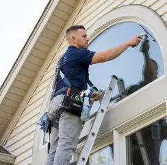 Professional window cleaner on a ladder using a squeegee to clean an exterior arched window, San Diego, CA.
